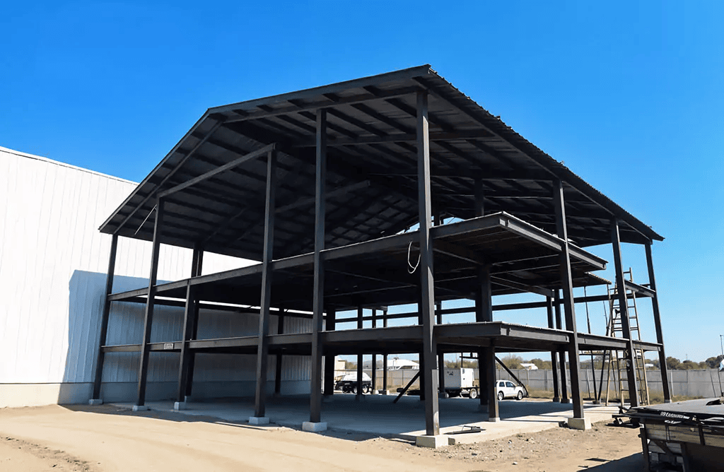 Pre-engineered steel building under construction with structural steel columns, beams, roofing framework and partially enclosed walls under a clear blue sky.